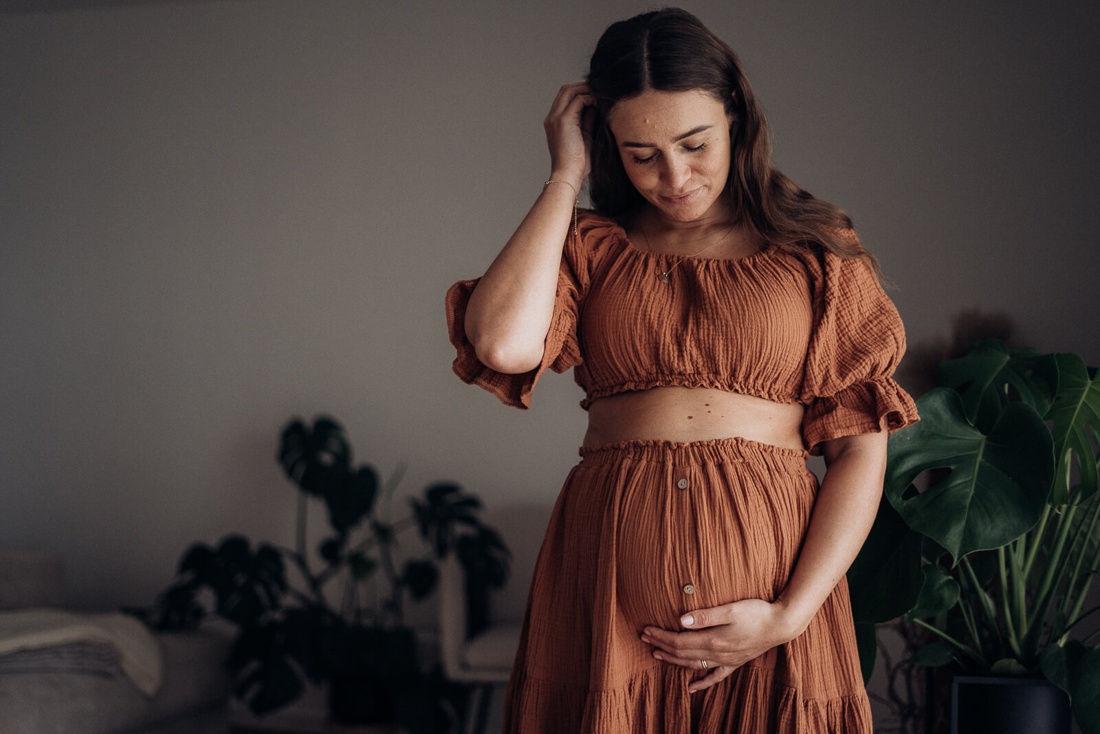 Schwangere steht in einem schönen Kleid im Wohnzimmer und streicht sich mit einer Hand liebevoll durchs Haar, während sie die andere Hand sanft um den Babybauch legt.