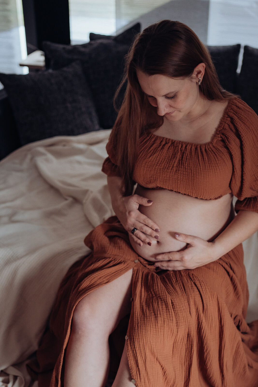 Werdende Mama sitzt in einem hellbraunen Kleid bei sich zuhaue auf der Couch und streichelt zärtlich ihren nackten Babybauch, während sie liebevoll zu ihrem Babybauch hinunter schaut.
