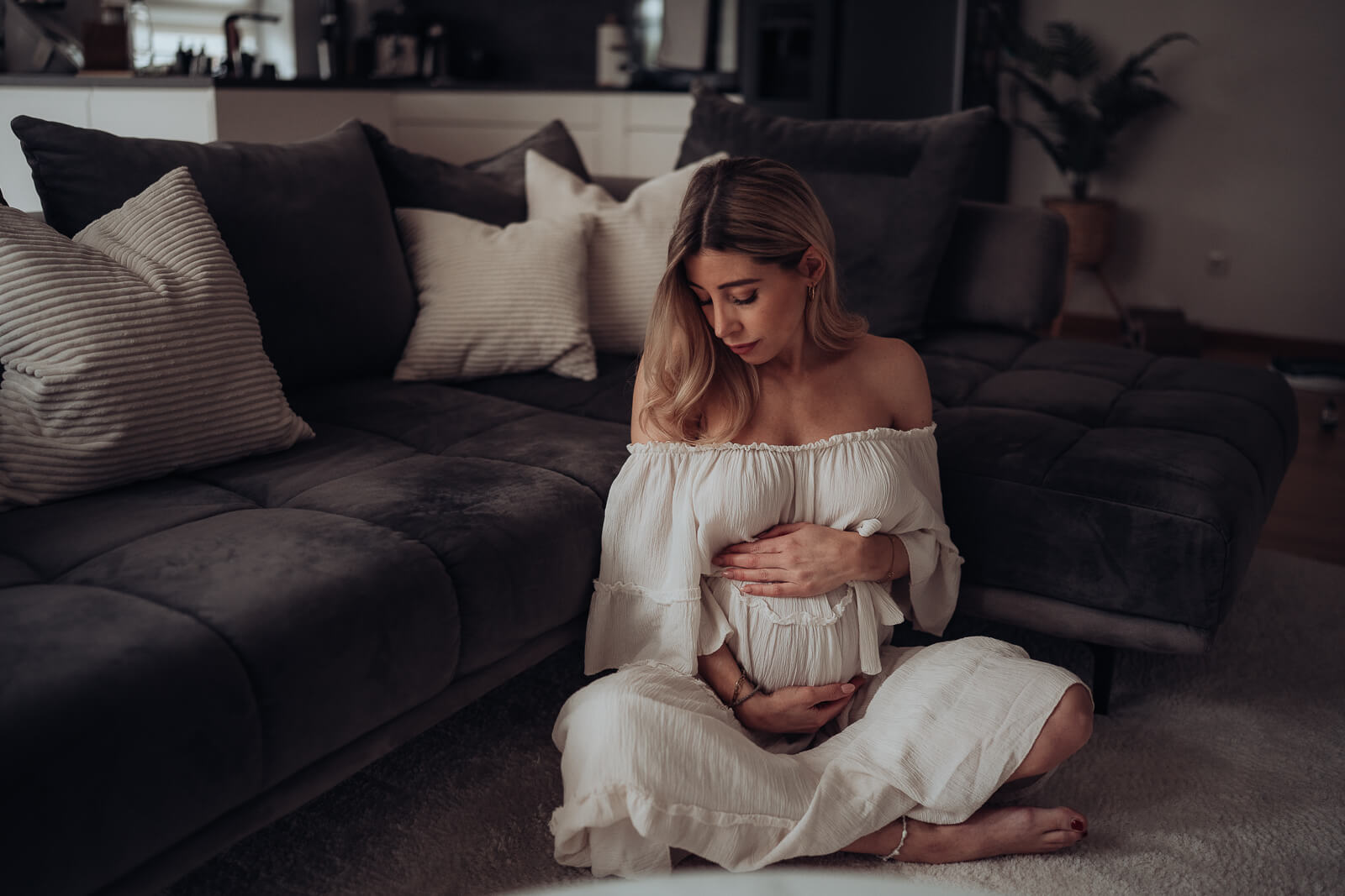 Babybauchshooting zuhause, bei dem die werdende Mama in einem schönen weißen Kleid auf dem Boden vor der Couch im Schneidersitz sitzt und ihren Babybauch mit beiden Händen umfasst.