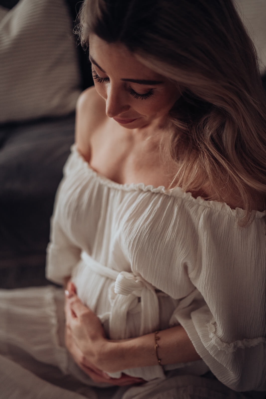 Nahaufnahme aus einem Babybauchshooting, bei dem die Schwangere im Schneidersitz vor der Couch sitzt und beide Hände sanft auf ihrem Babybauch liegen hat und hinunter schaut.