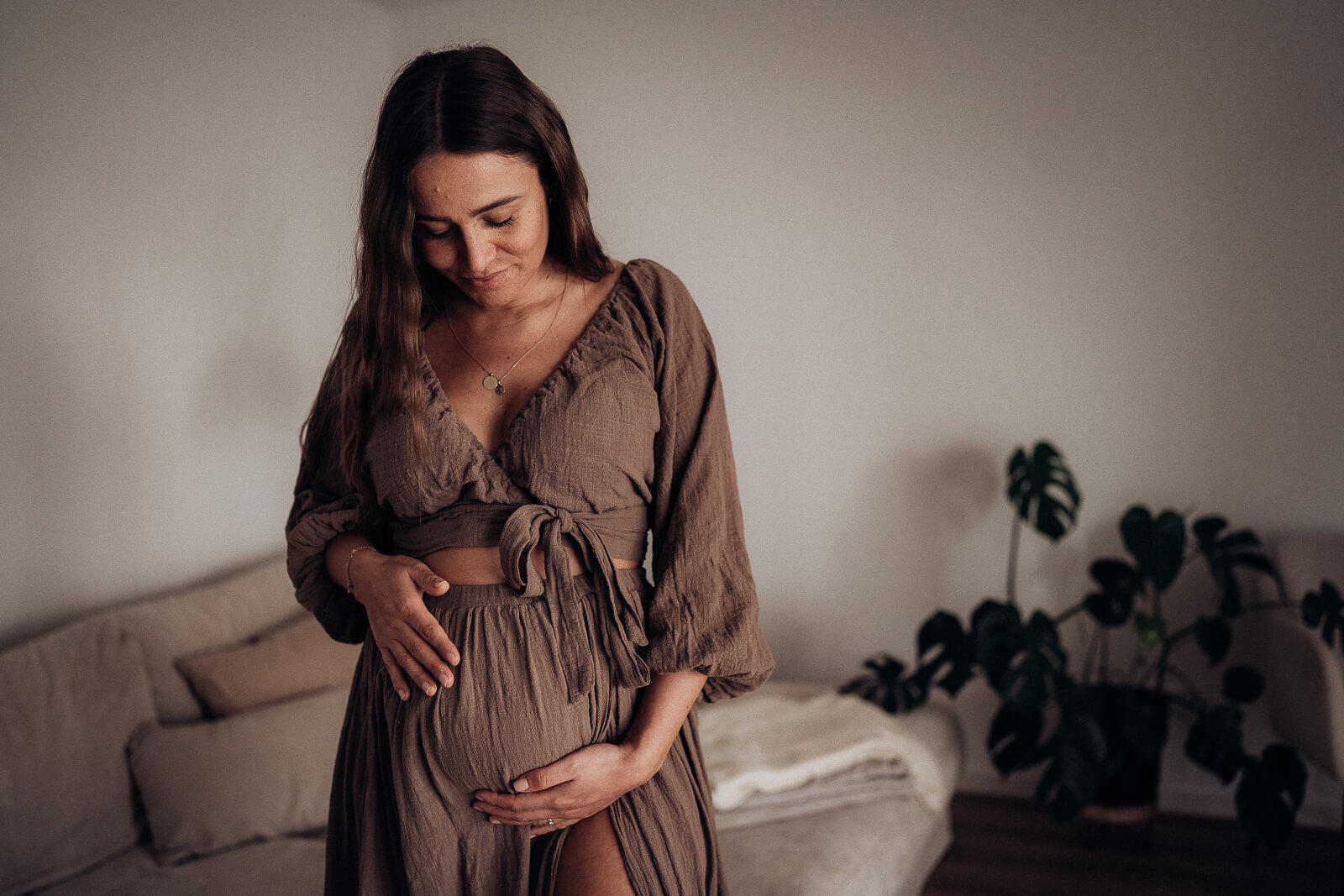 Schwangere steht in einem wunderschönen Kleid im Wohnzimmer und schaut vorfreudig zu ihrem Babybauch hinunter.