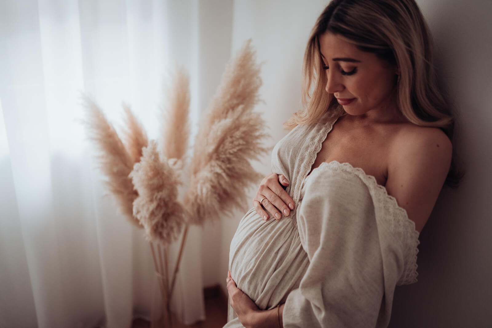 Schwanger steht an eine Wand gelehnt in einem wunderschönen hellen Kleid im Schlafzimmer und hält voller Liebe ihren Babybauch.