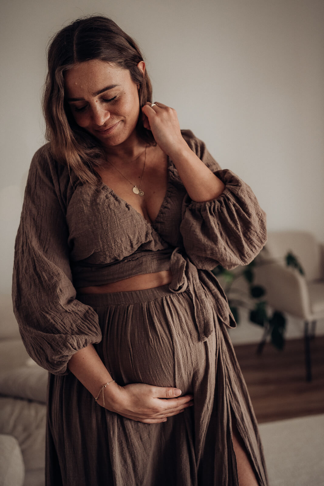 Eine Schwangere steht mit einem wunderschönen braunen Kleid in ihrem Wohnzimmer und streicht sich mit einer Hand die Haare hinters Ohr, dabei hält die andere Hand liebevoll den Babybauch.