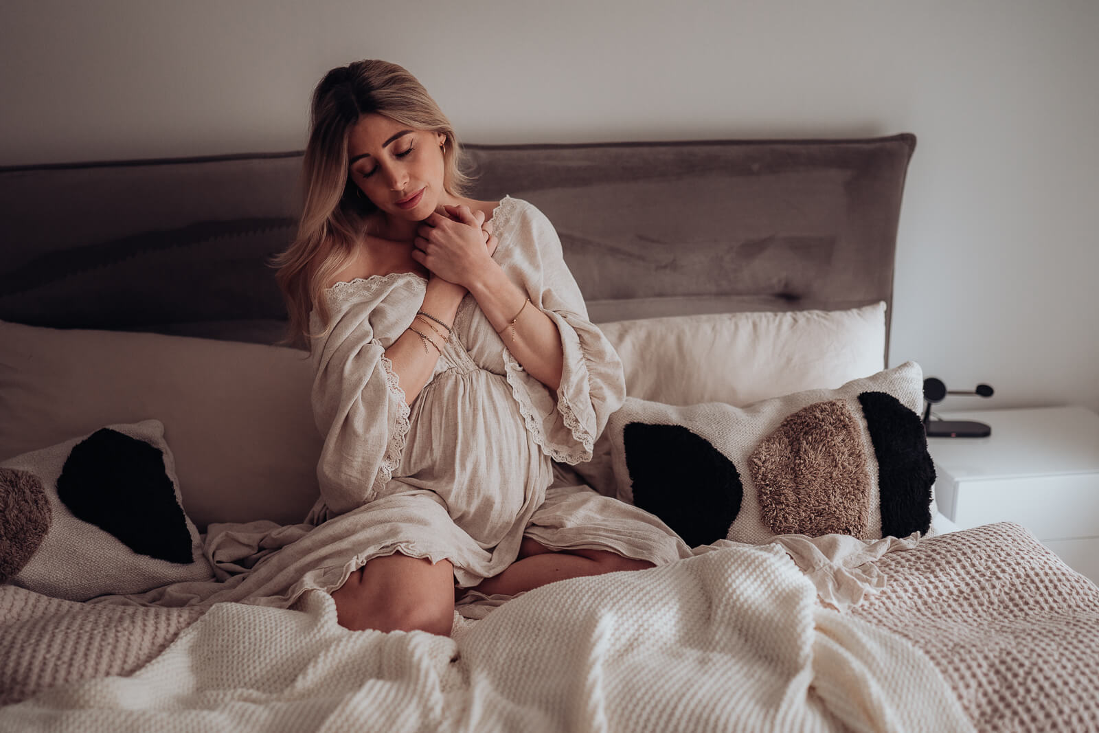Babybauchshooting Zuhause bei dem die Schwangere auf dem Bett sitzt und beide Hände auf ihrer Brust ablegt und mit geschlossenen Augen den Moment genießt.