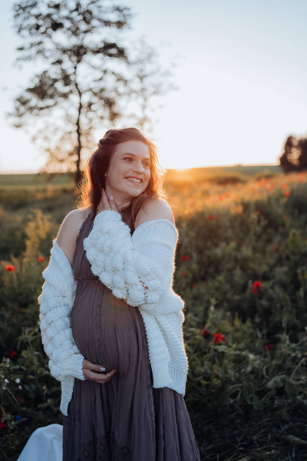 Schwangere steht mit einem schönen Kleid und einer gemütlichen Strickweste in einem Mohnblumenfeld und streichelt sich selbst am Hals, während sie mit der anderen Hand liebevoll ihren Babybauch hält.