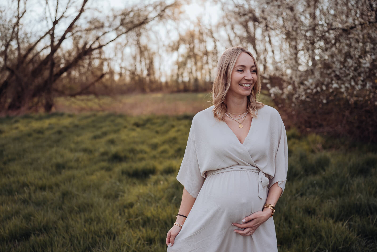 Schwangere in schönem beigefarbenem Kleid steht auf der Wiese und schwingt sanft das Kleid, während sie mit der anderen Hand liebevoll  ihren Babybauch hält.