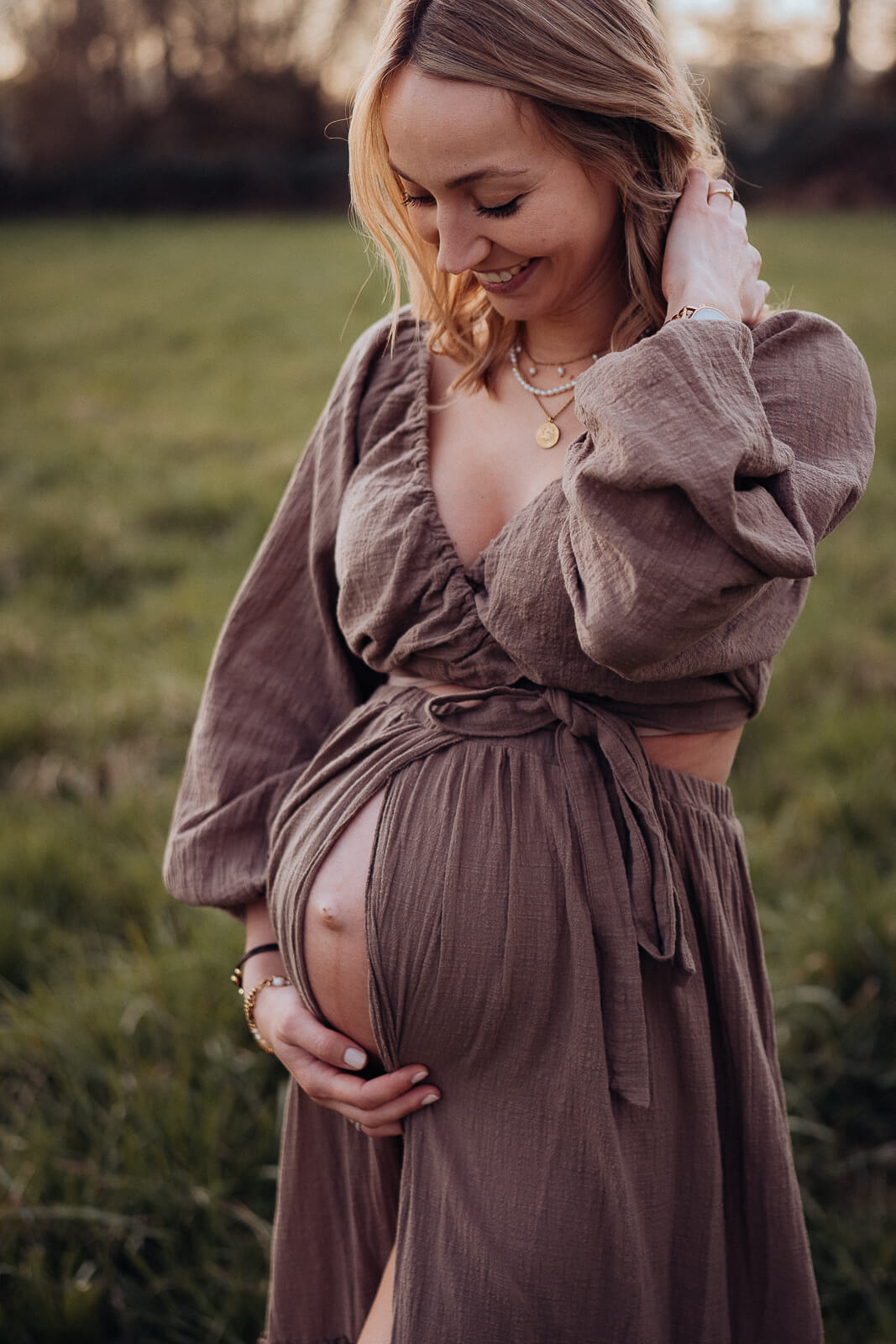 Babybauchsooting auf einer Wiese mit einer Schwangeren in einem braunen Kleid, der Babybauch spitzelt ein wenig aus dem Kleid hervor und die werdende Mama streicht sich lächelnd die Haare hinters Ohr.
