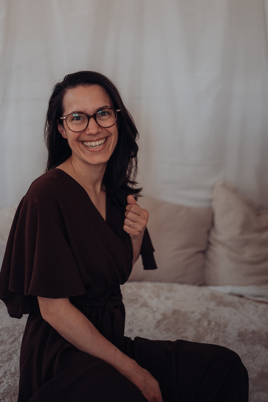 Babybauch und Motherhood Fotografin Sarah Bomm sitzt bei einer Homestory in einem braunen Kleid seitlich auf dem Boden und lächelt in die Kamera.