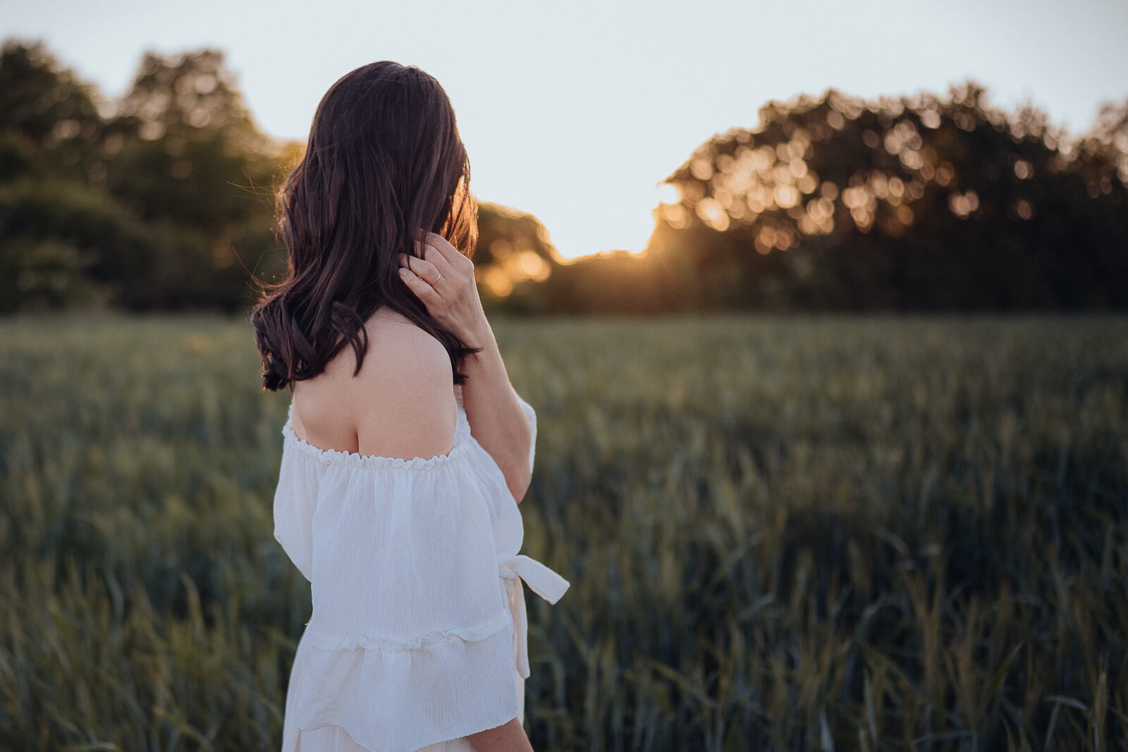 Babybauch und Motherhood Fotografin Sarah Bomm steht mit einem weißen Kleid zur Sonne im Hintergrund gewendet vor einem Weizenfeld und streicht sich durchs Haar.