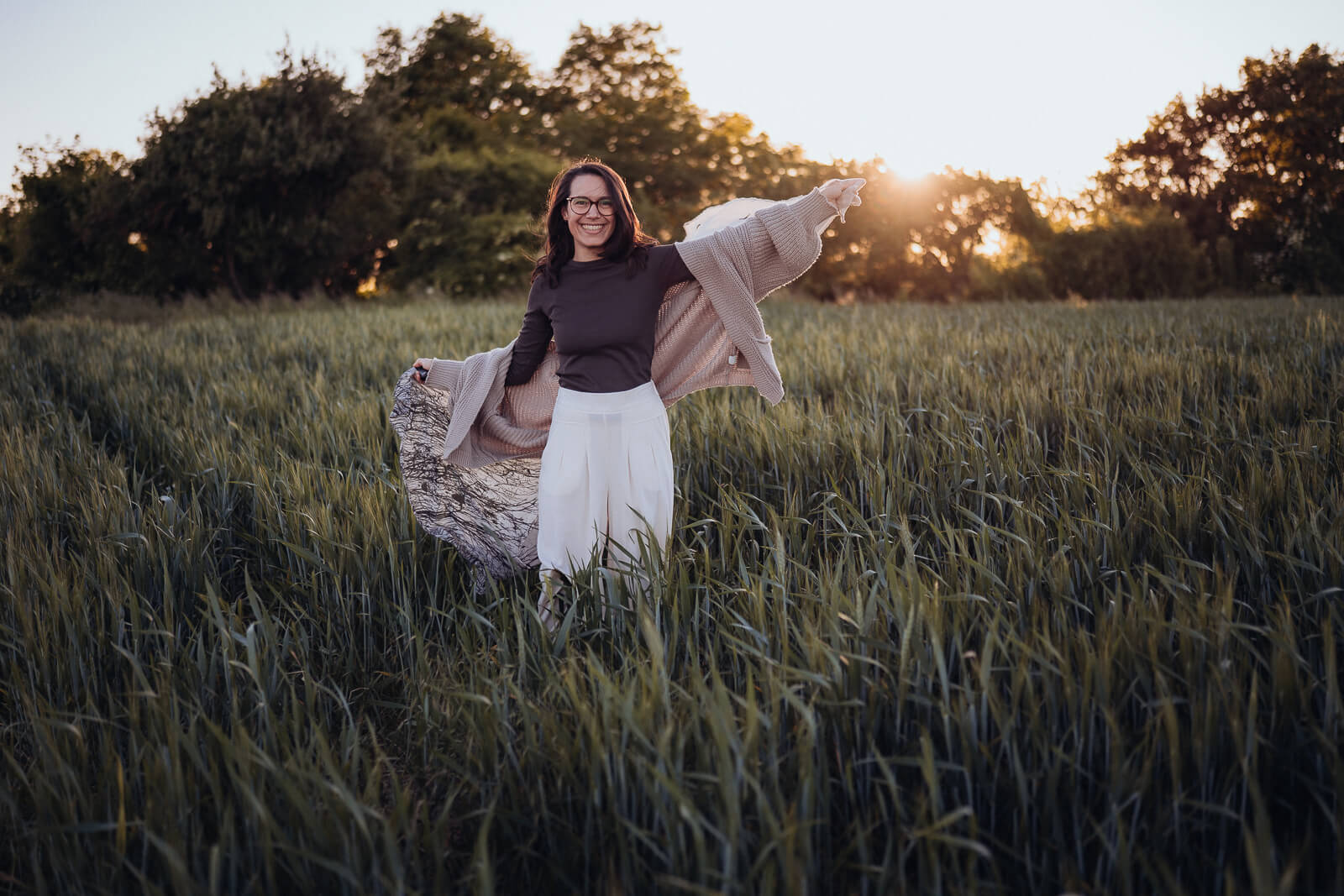 Babybauch und Motherhood Fotografin Sarah Bomm steht in einem grünen Weizenfeld mit dem Sonnenuntergang im Rücken und schaut lächelnd in die Kamera, dabei schwingt sie einen luftigen Schal hinter sich.