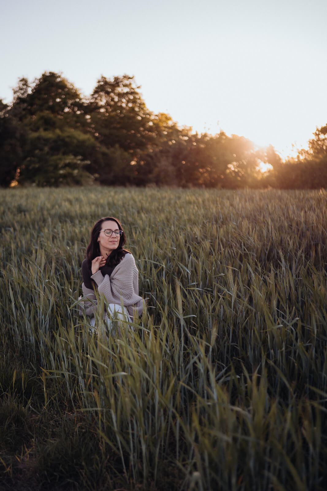 Babybauch und Motherhood Fotografin Sarah Bomm kniet bei Sonnenuntergang im grünen Weizenfeld mit geschlossenen Augen und spielt in ihren Haaren.