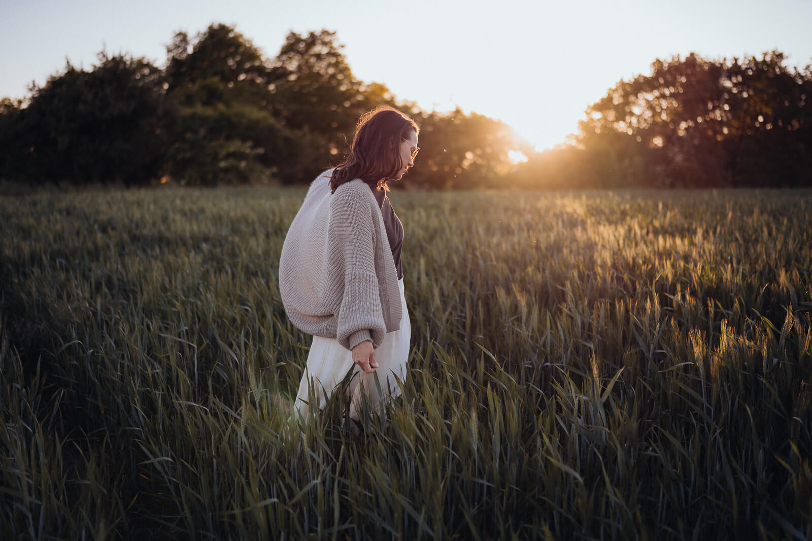Babybauch und Motherhood Fotografin Sarah Bomm steht seitlich in grünem Weizenfeld mit der untergehenden Sonne im Hintergrund und sie streift mit ihrer Hand sanft durch das Weizen.