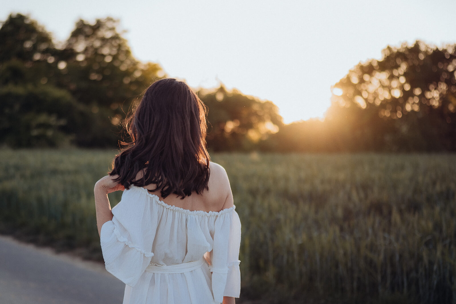 Babybauch und Motherhood Fotografin Sarah Bomm steht zur Sonne gewandt vor einem Weizenfeld in einem weißen Kleid und spielt in ihren Haaren.