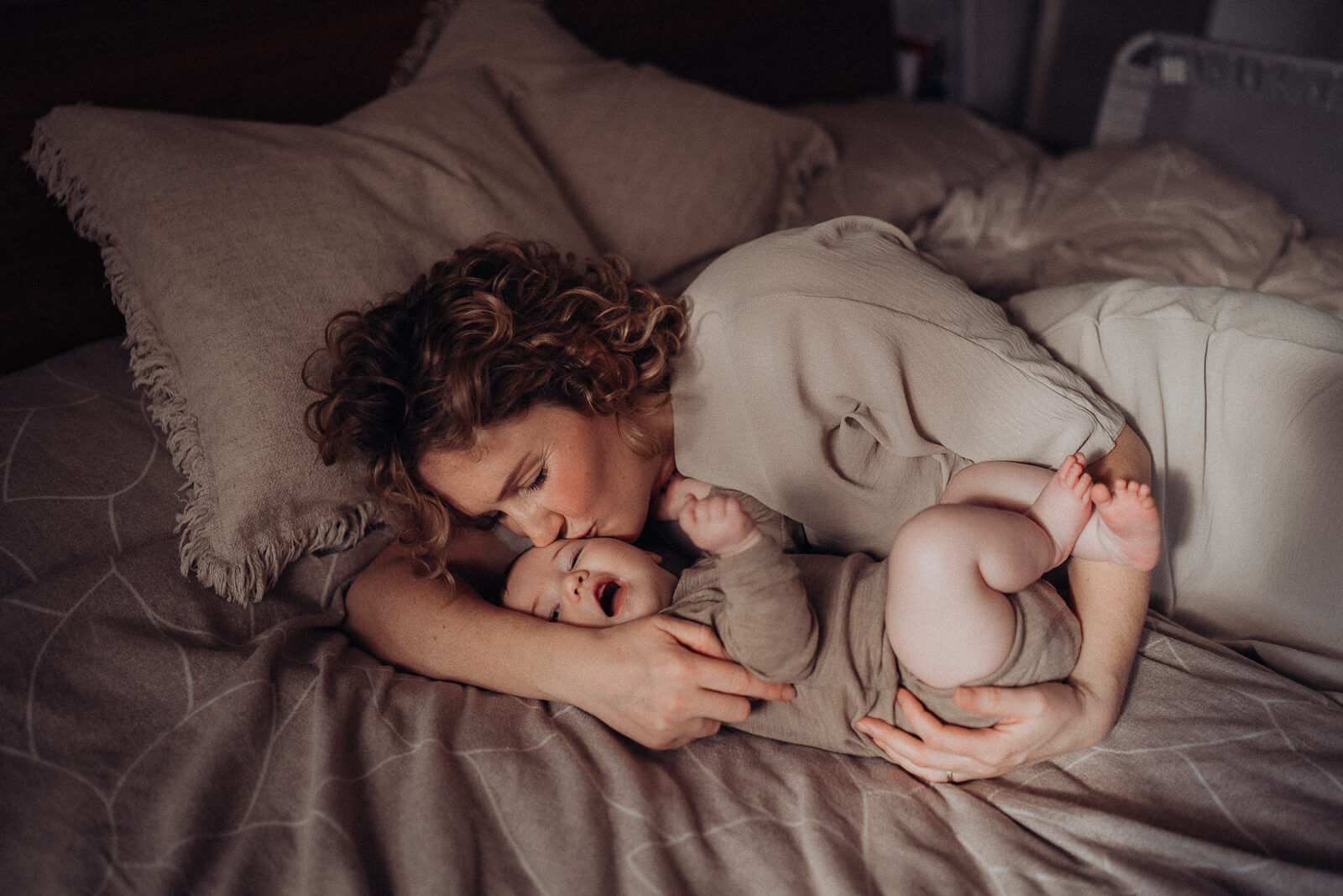 Mama kuschelt sich im Bett liegend an ihr Baby und gibt ihm sanft einen Kuss auf die Schläfe.