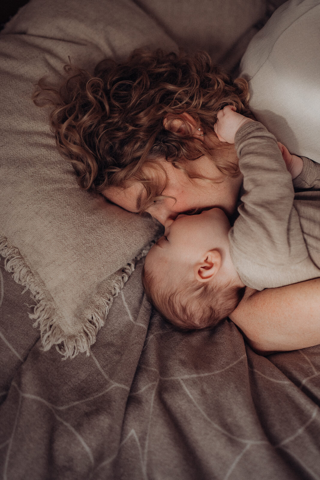 Kuscheliger Motherhood Moment zwischen Mama und Baby im Bett, während das Baby der Mama in den Haaren spielt und sein Gesicht ganz nah an das der Mama drückt.