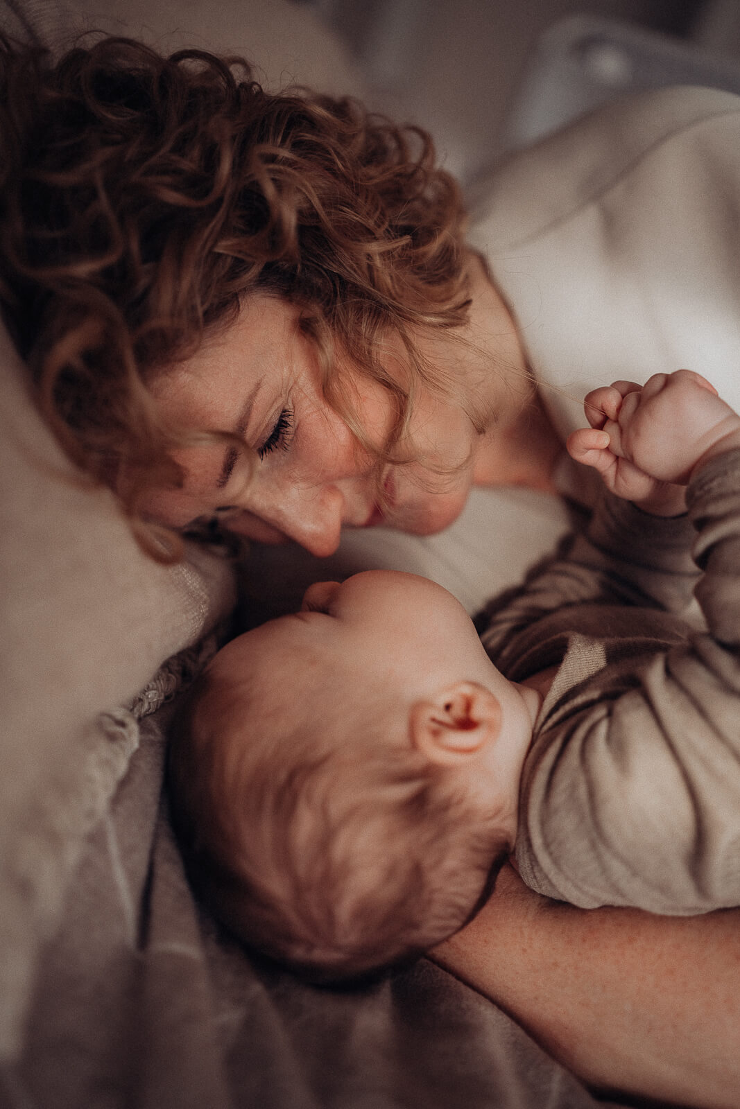 Motherhood-Kuschelmoment einer Mama, die mit ihrem Baby im Arm auf dem Bett liegt und sich liebevoll anschauen.