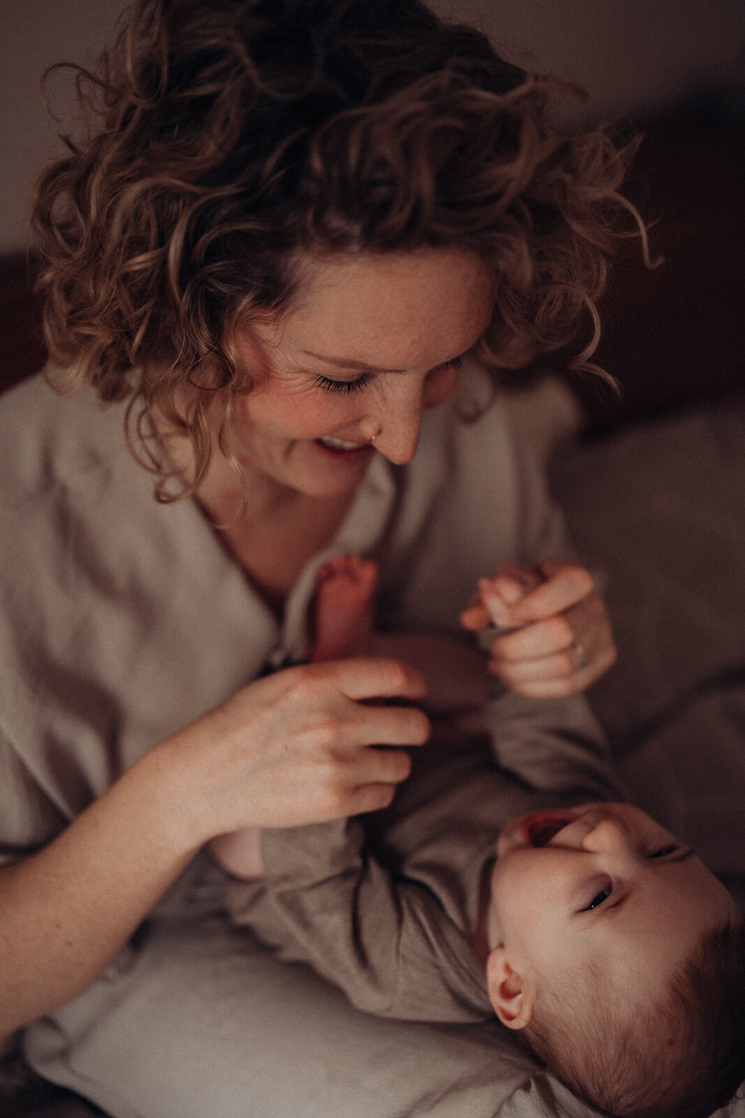 Mama sitzt im Bett und hat ihr Baby im Schoß liegen, es schaut sie an und beide Lachen.