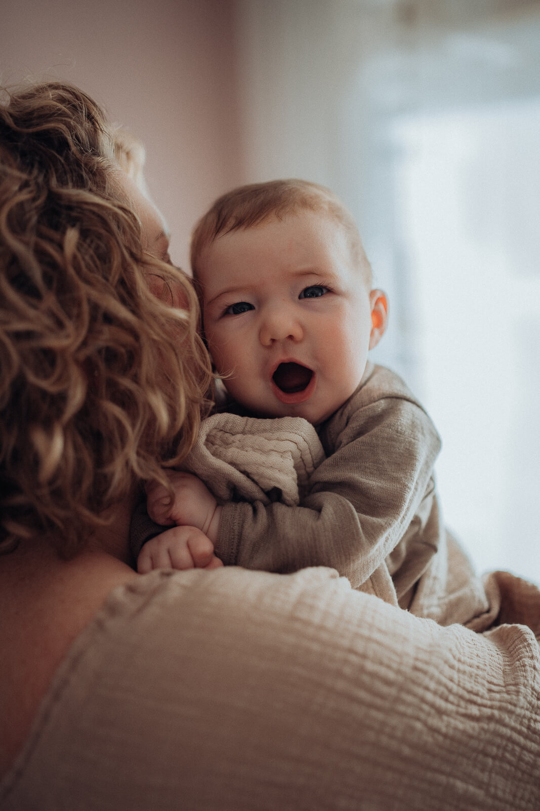 Baby schaut während eines Mommy and me Shootings über die Schulter der Mama, die entspannt zuhause in ihrem Wohnzimmer steht.