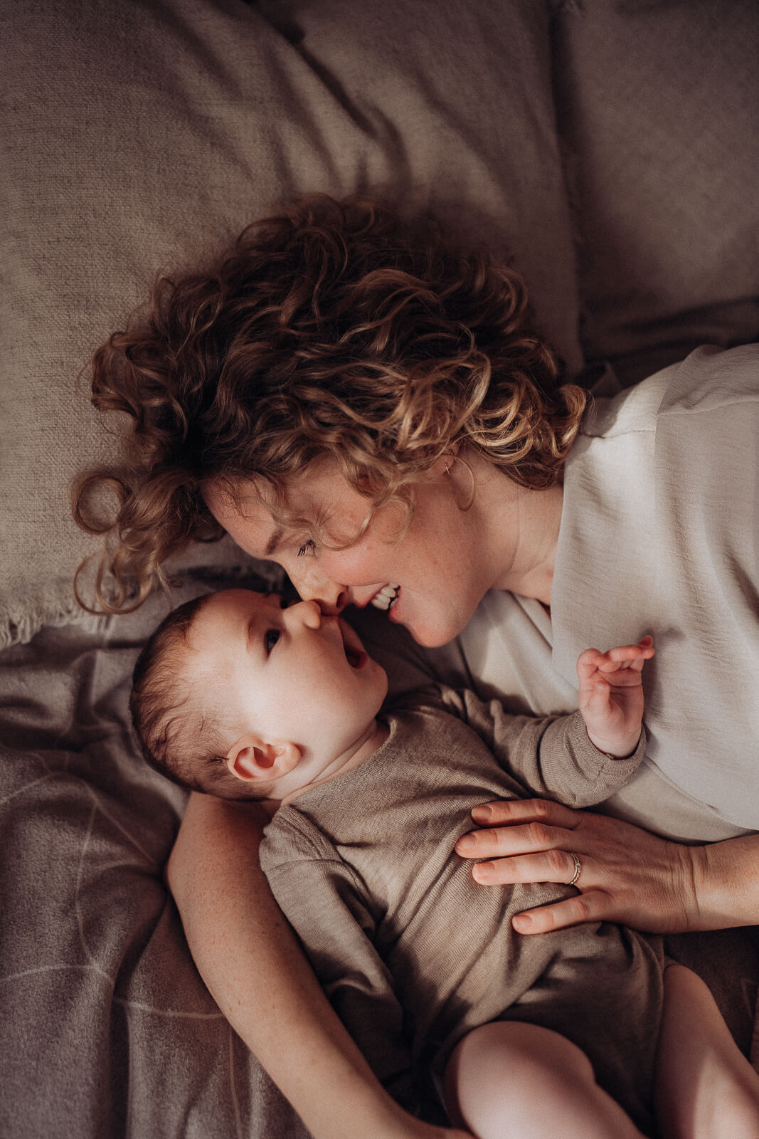 Mama liegt mit ihrem Baby kuschelnd im Bett und gibt ihm einen Nasenkuss.